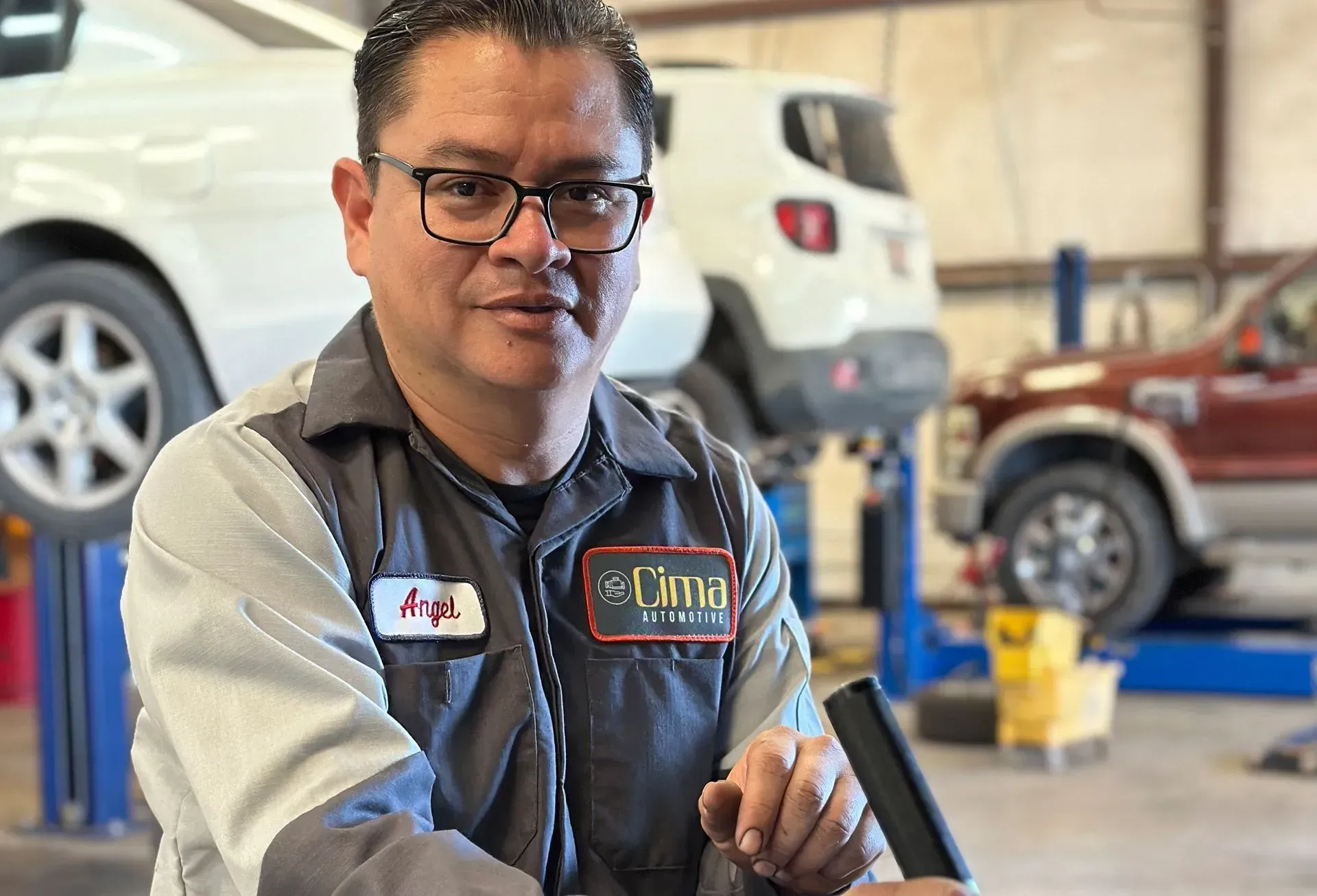 Mechanic in grey uniform, glasses, in a garage. Cars on lifts in background.