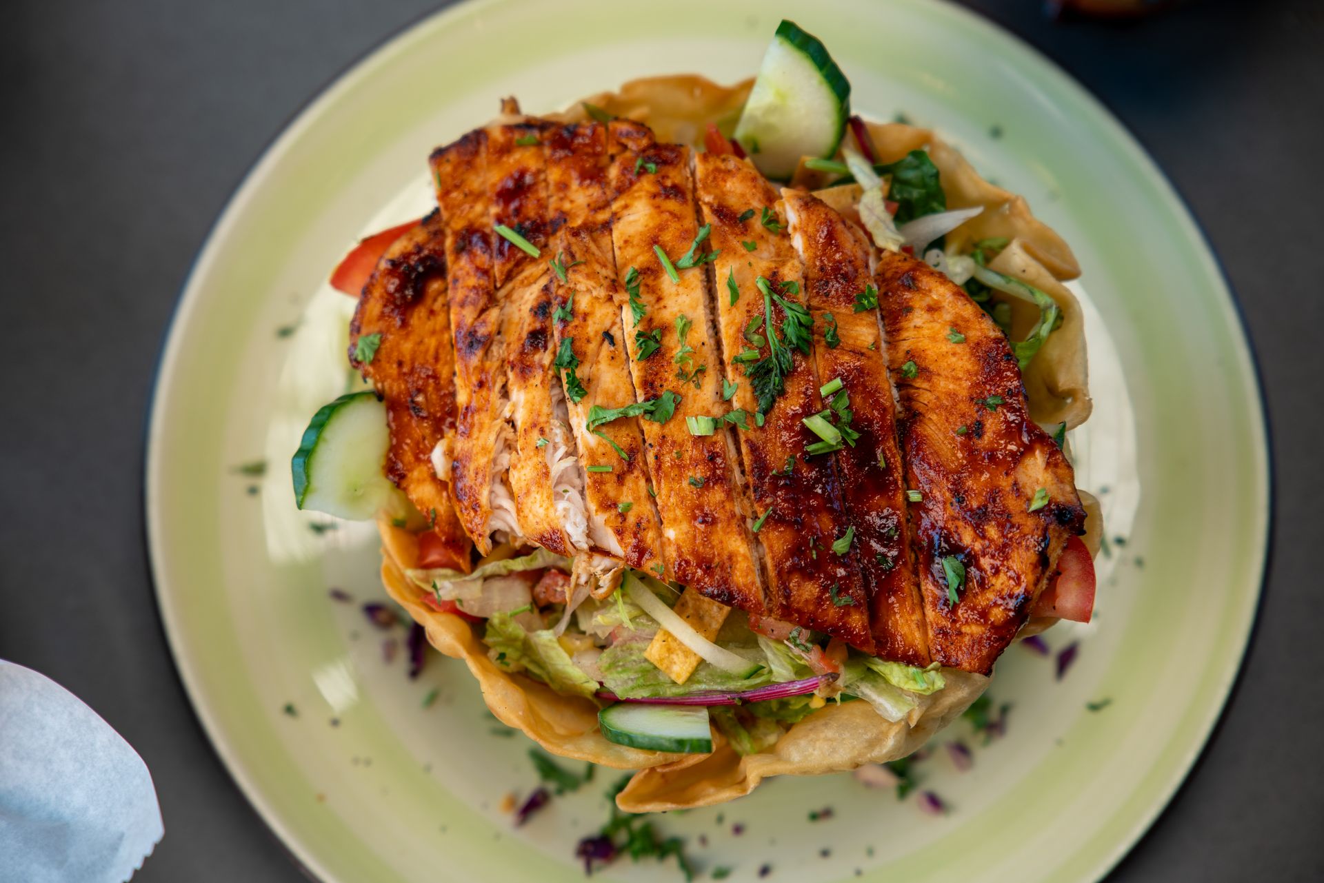 Grilled chicken salad in a fried tortilla bowl, with cucumber and lettuce on a green plate.