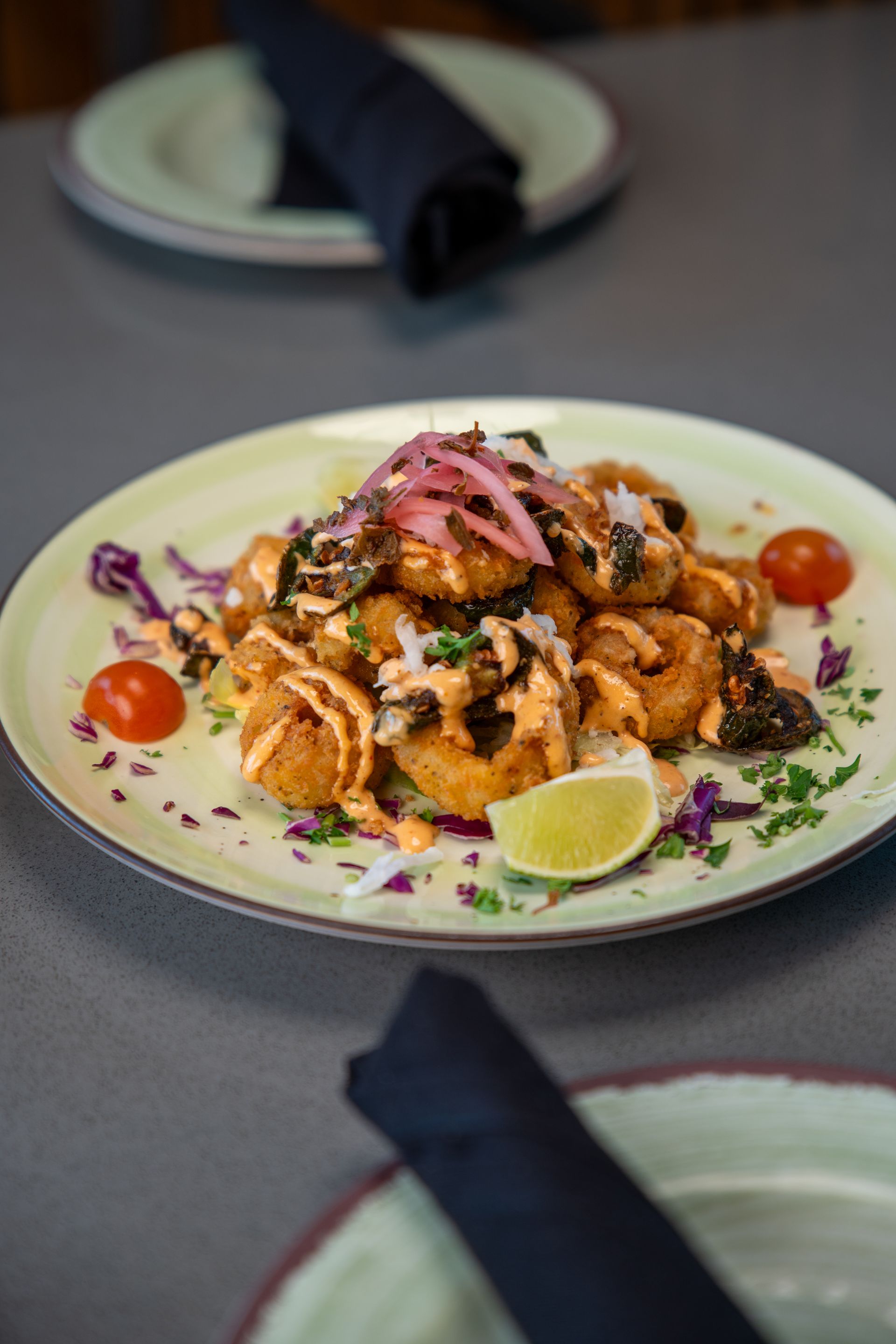 Plate of fried calamari with sauce, lime, tomatoes, and pickled onions on a restaurant table.