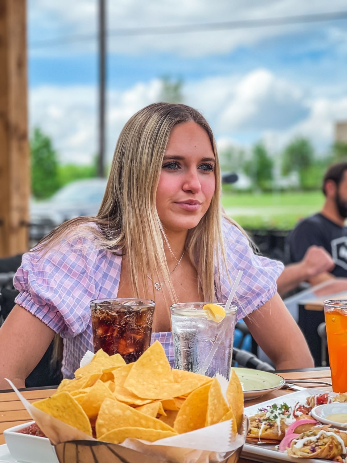 Woman at a restaurant table with chips, drinks, and food. She gazes to the side. Outdoor setting.