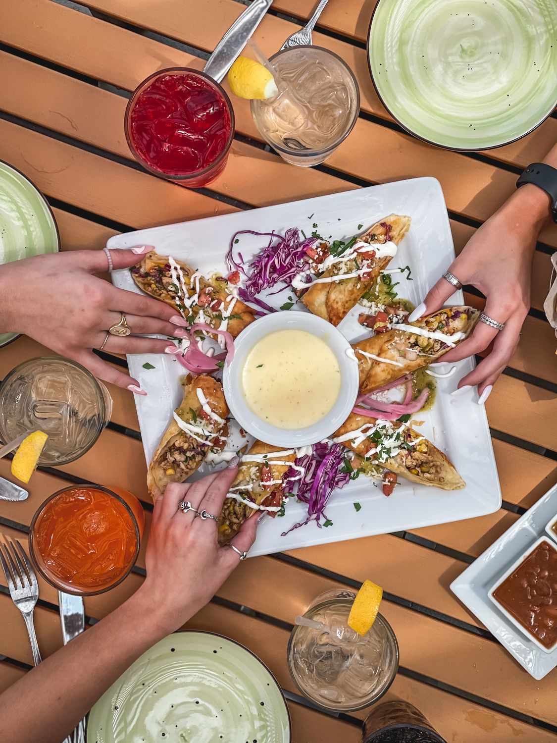 People sharing a plate of food, surrounded by drinks, on a wooden table.