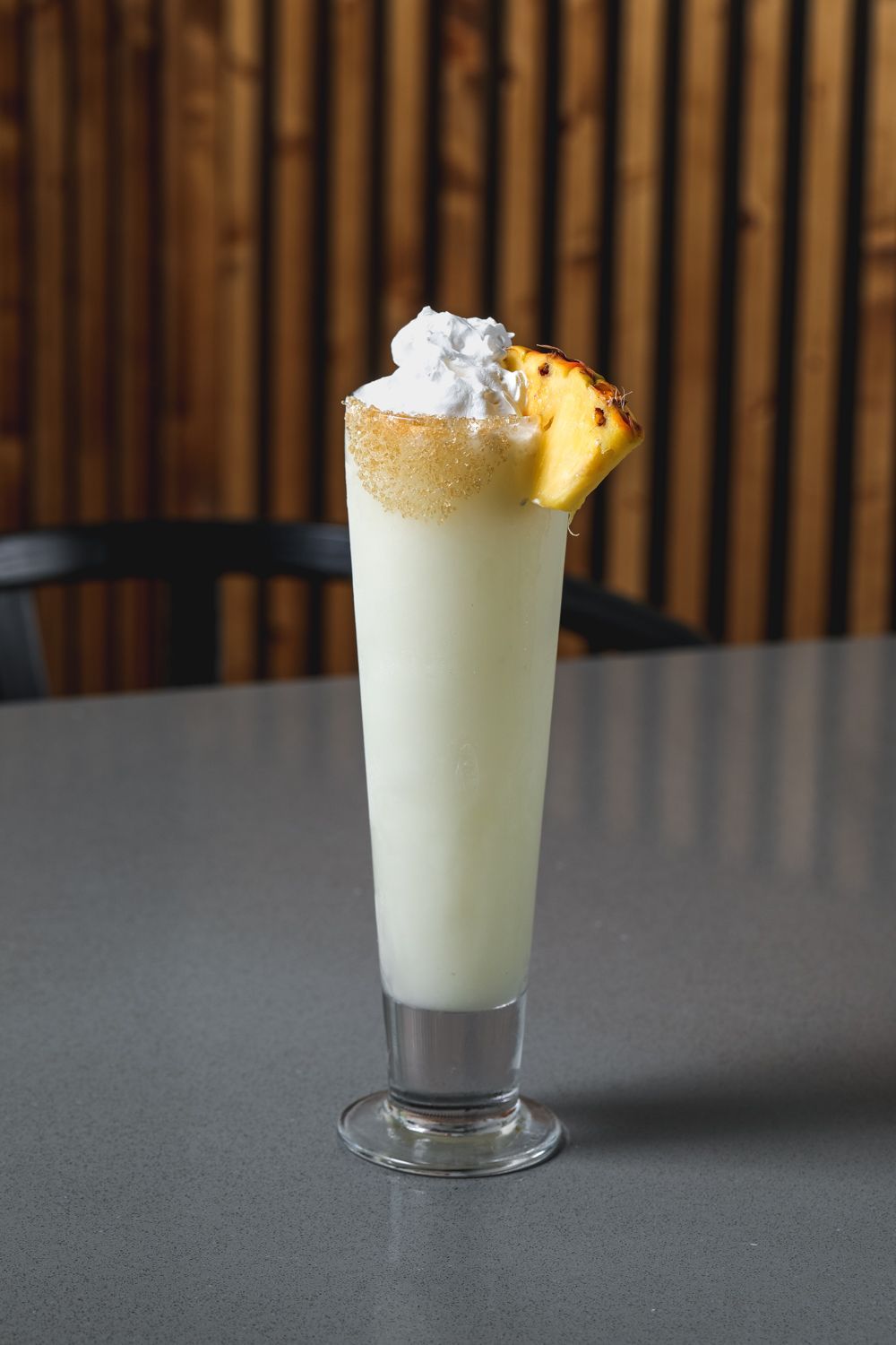 Tall glass of pale drink with whipped cream and pineapple garnish on a gray surface, wooden background.