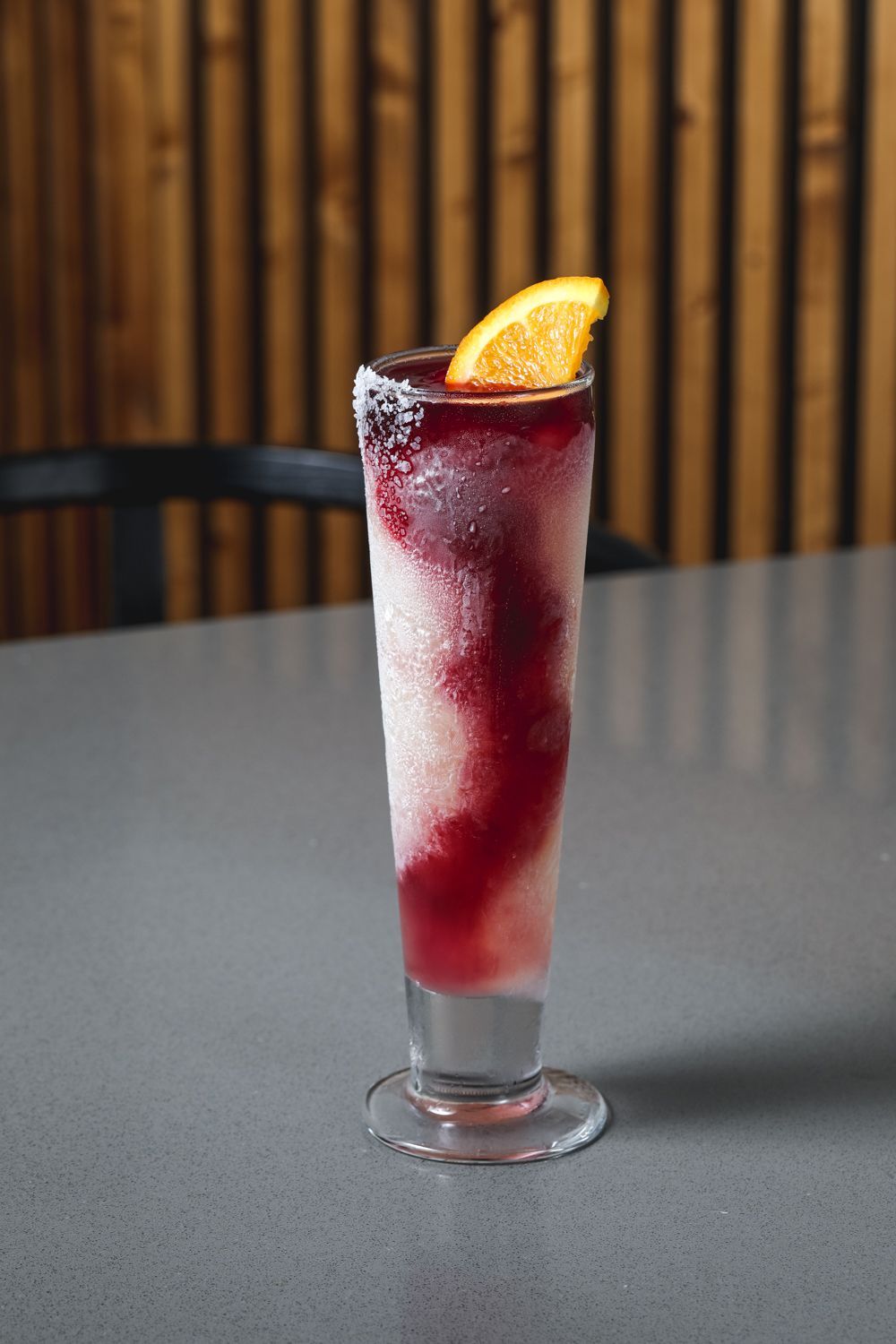 Tall, frosted cocktail with a dark red layer topped with an orange slice, on a gray surface against a wood-paneled wall.
