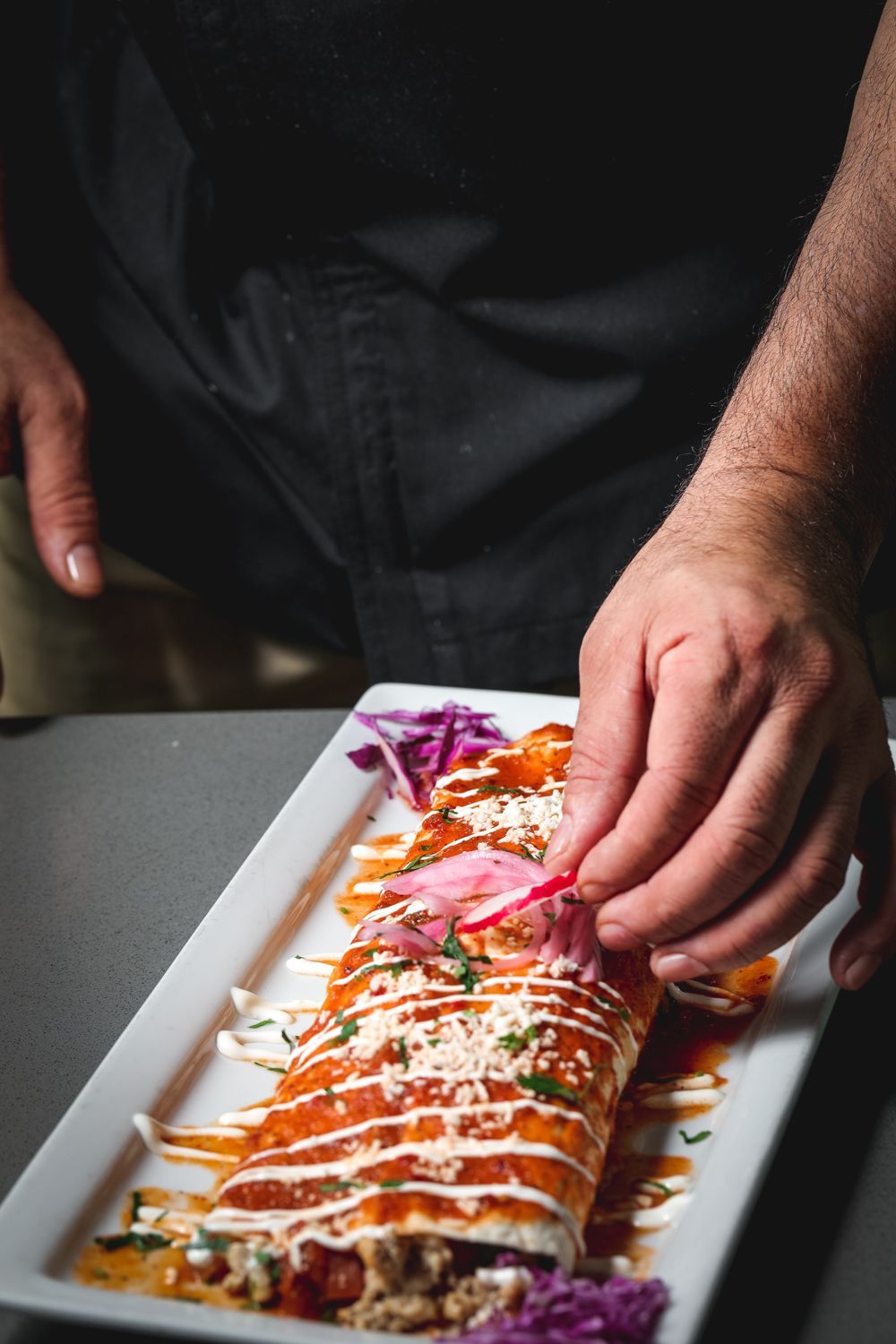 Chef garnishes a plated enchilada with pink pickled onions.