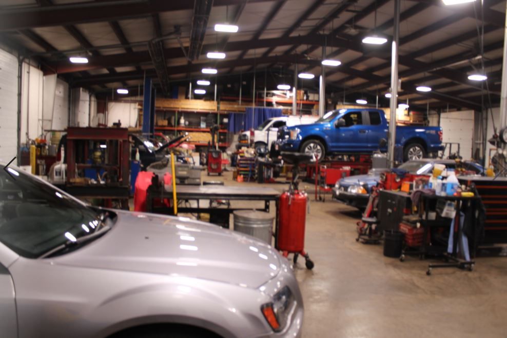 Interior of a car repair shop. Several vehicles are being worked on, including a blue pickup truck on a lift. Tools and equipment are scattered around.