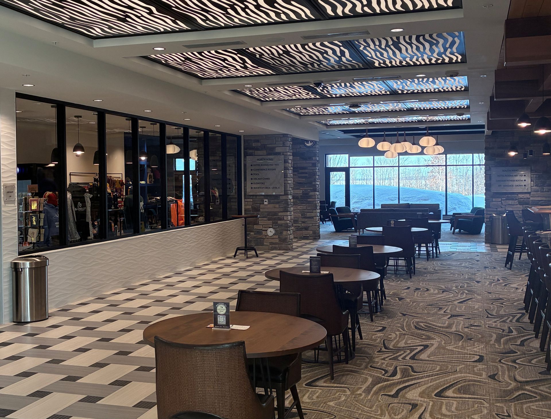 A brightly lit lounge area with round tables, chairs, and patterned ceilings, featuring a glass-walled gift shop nearby.