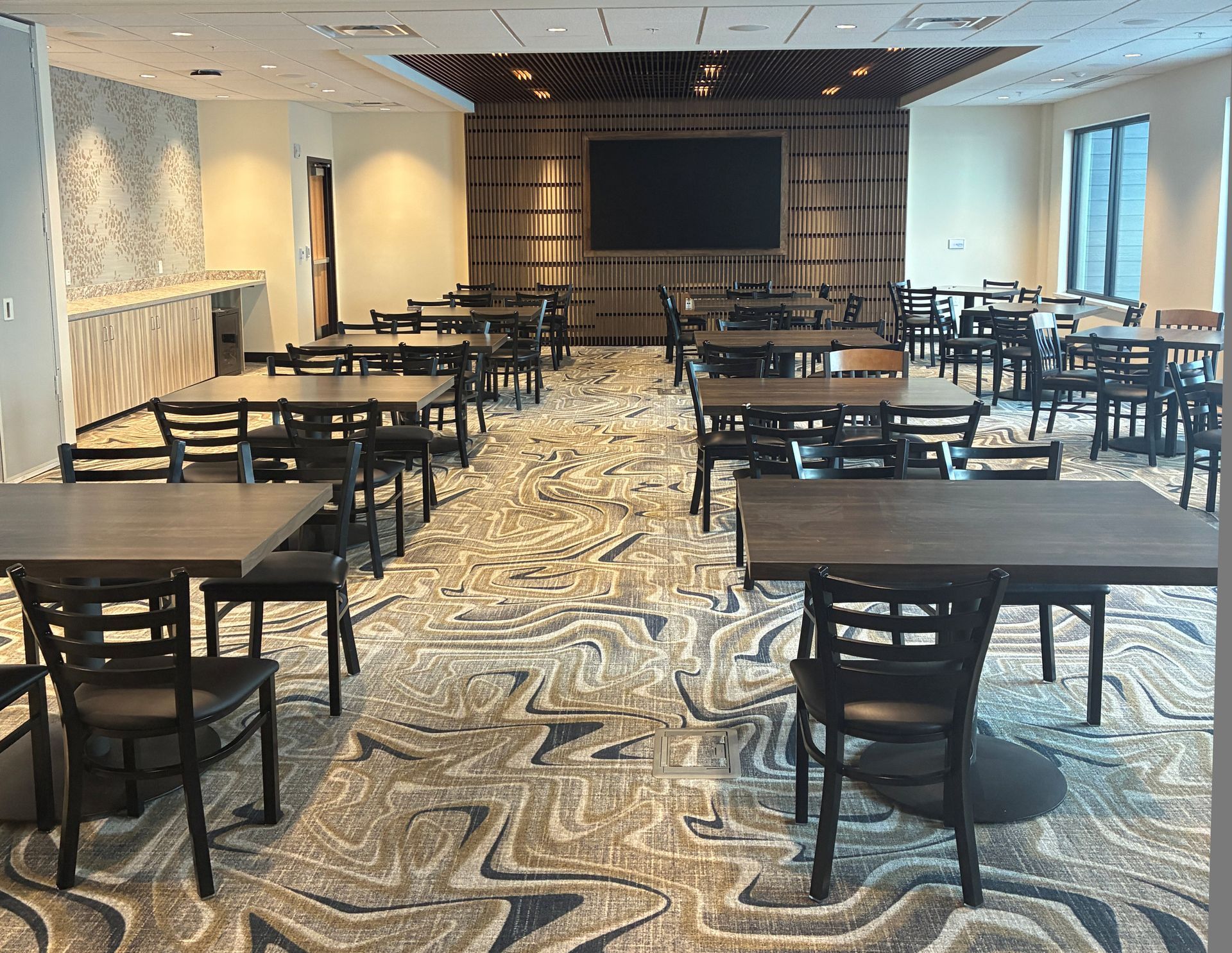 A room with multiple dark wood dining tables and chairs arranged on a patterned carpet under recessed lighting.