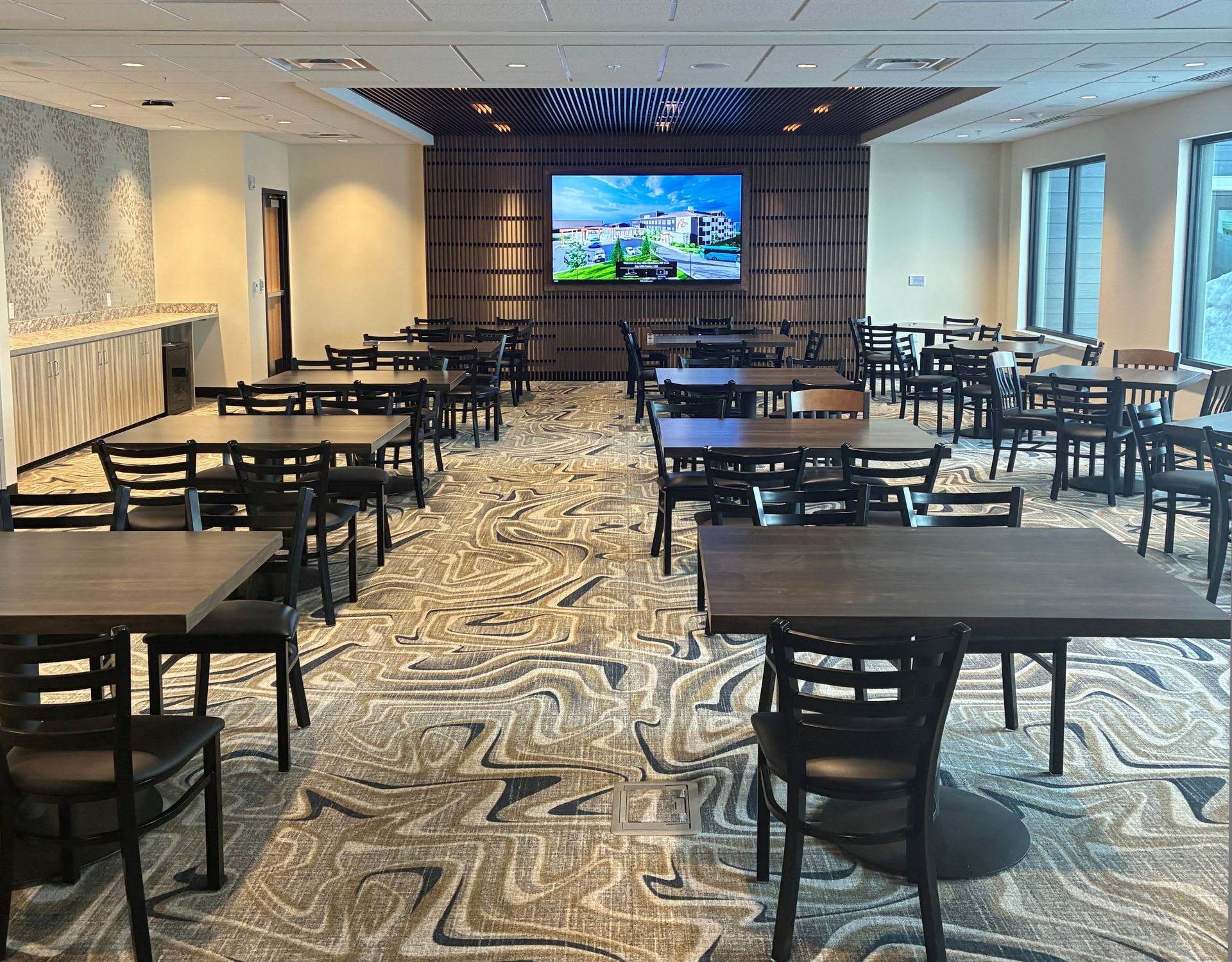 A dining room with many square wooden tables and black chairs arranged in rows on a patterned grey and white carpet.