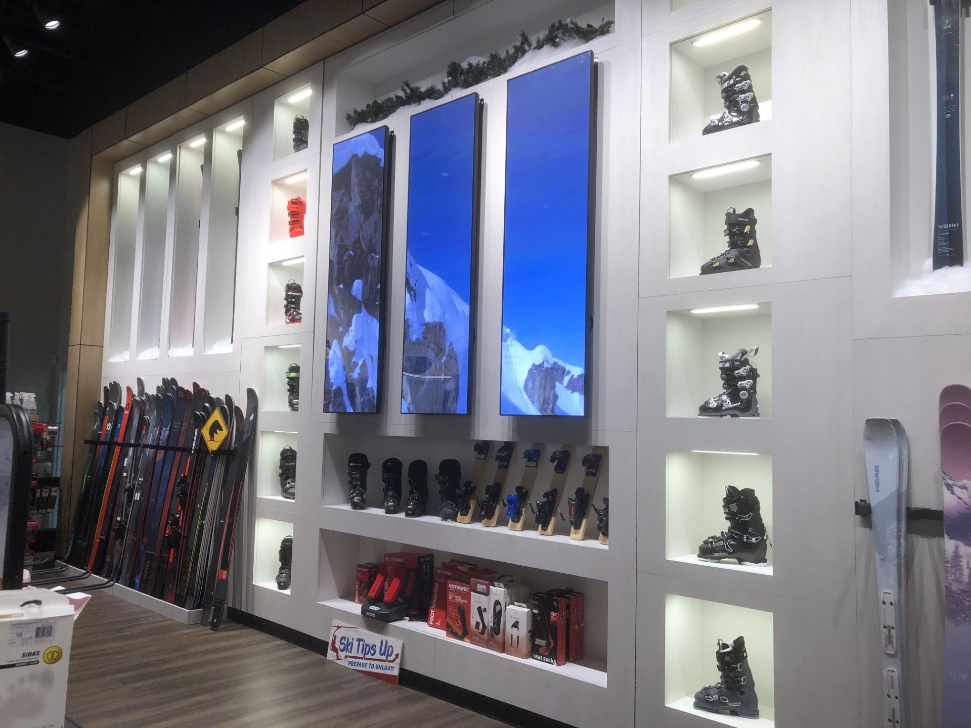 A store display wall featuring rows of alpine ski boots, skis, and three large vertical digital screens showing snow scenes.