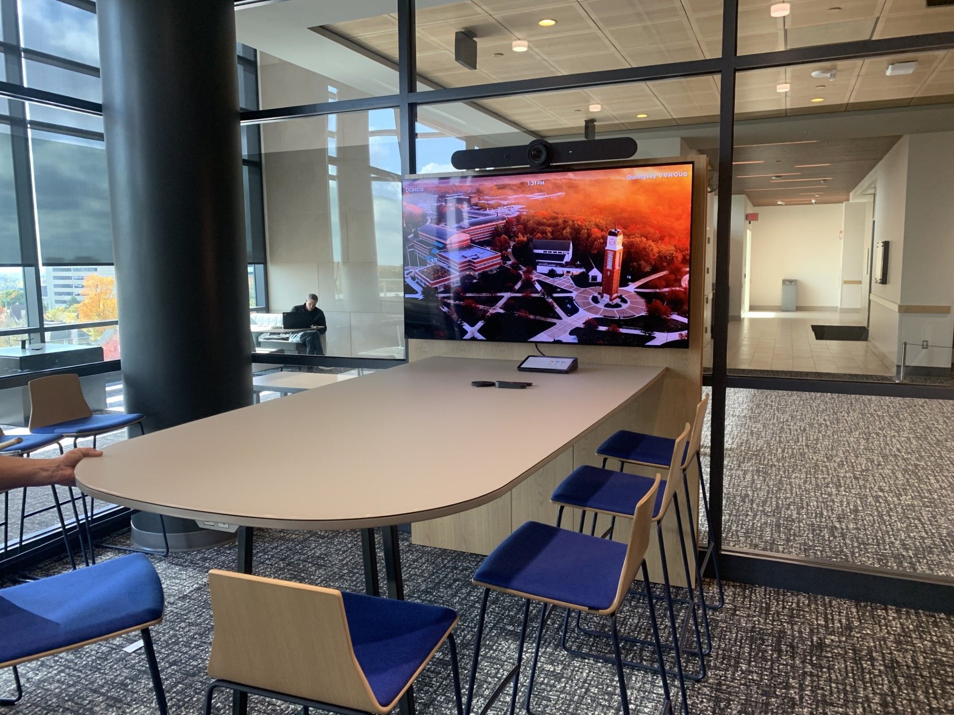 A conference table with blue bar stools facing a wall-mounted monitor displaying a scenic landscape in an office space.