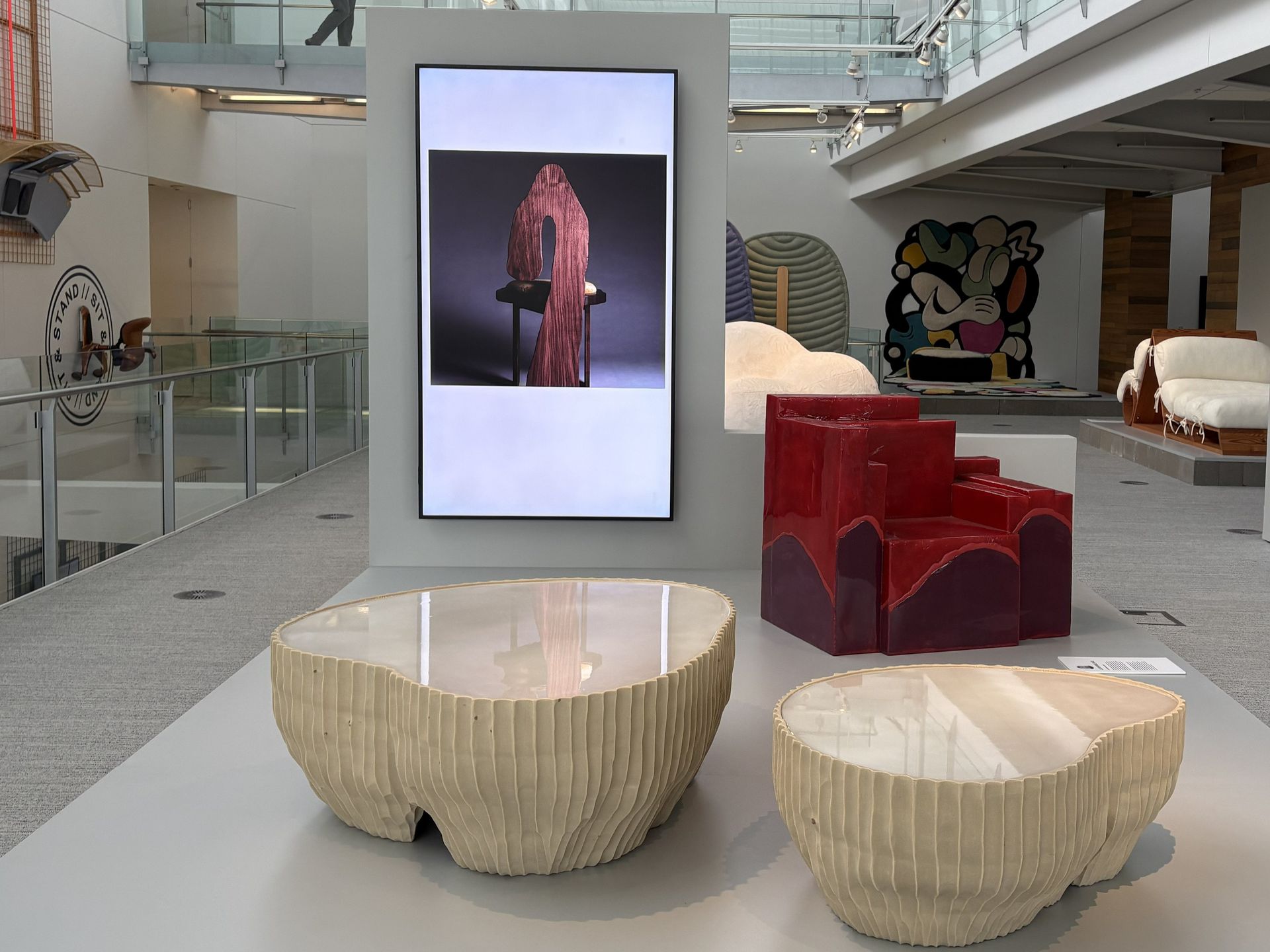 Modern furniture is exhibited in a gallery featuring two textured, beige coffee tables and a glossy red armchair.