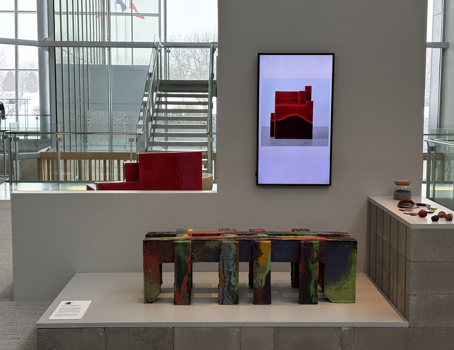 A multi-colored, rectangular, leg-based sculpture on a pedestal in a gallery near a monitor showing a red abstract form.