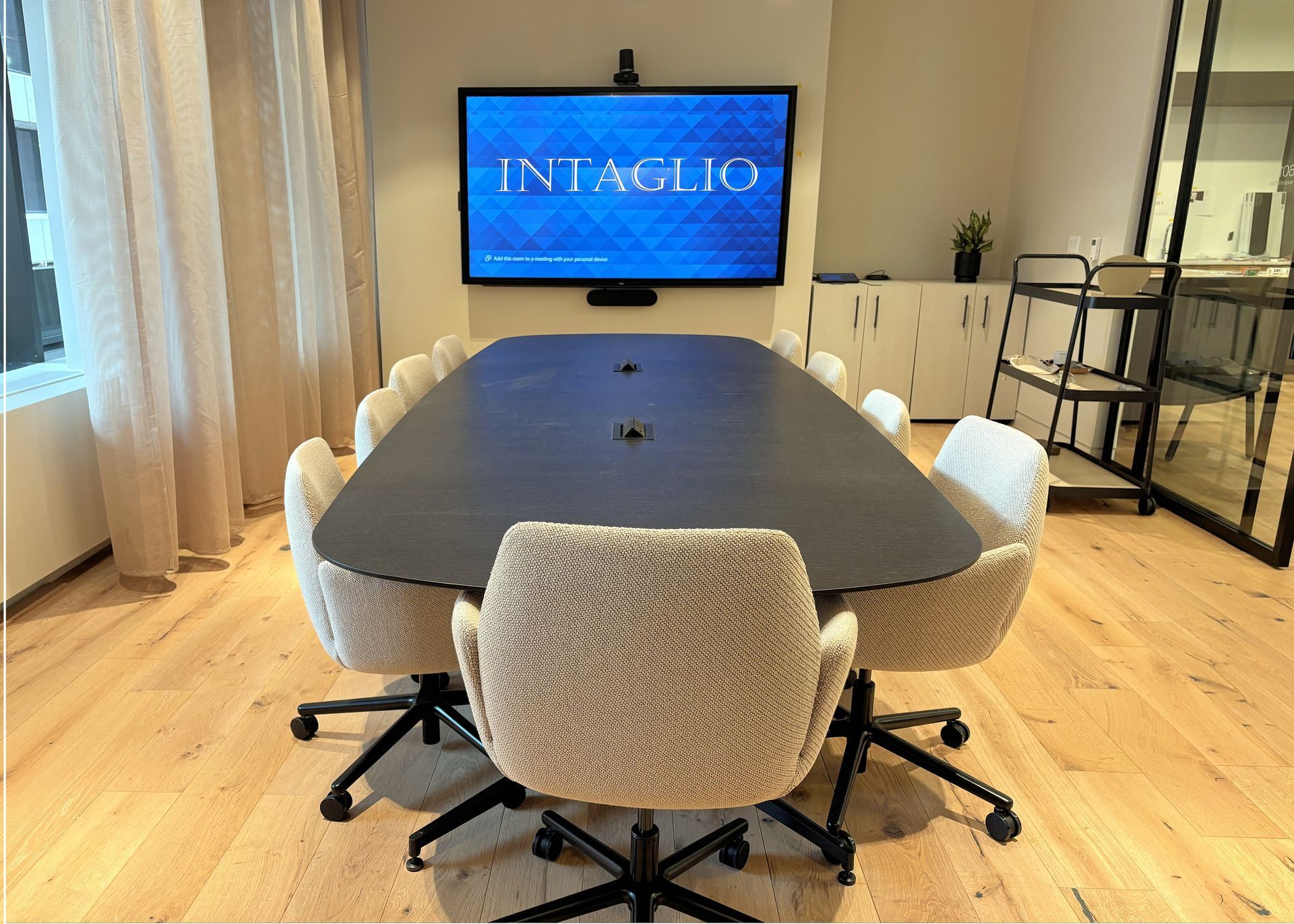 A conference room featuring a dark oval table surrounded by textured white chairs on light wood floors facing a TV screen.