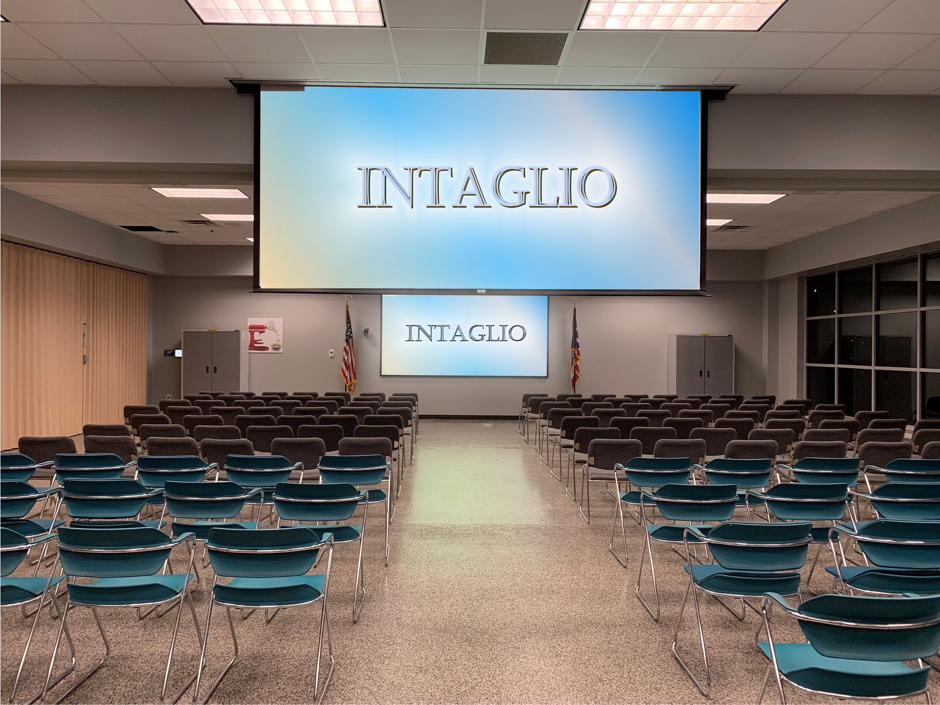 An empty conference room with rows of teal chairs facing a large projection screen displaying the word INTAGLIO.
