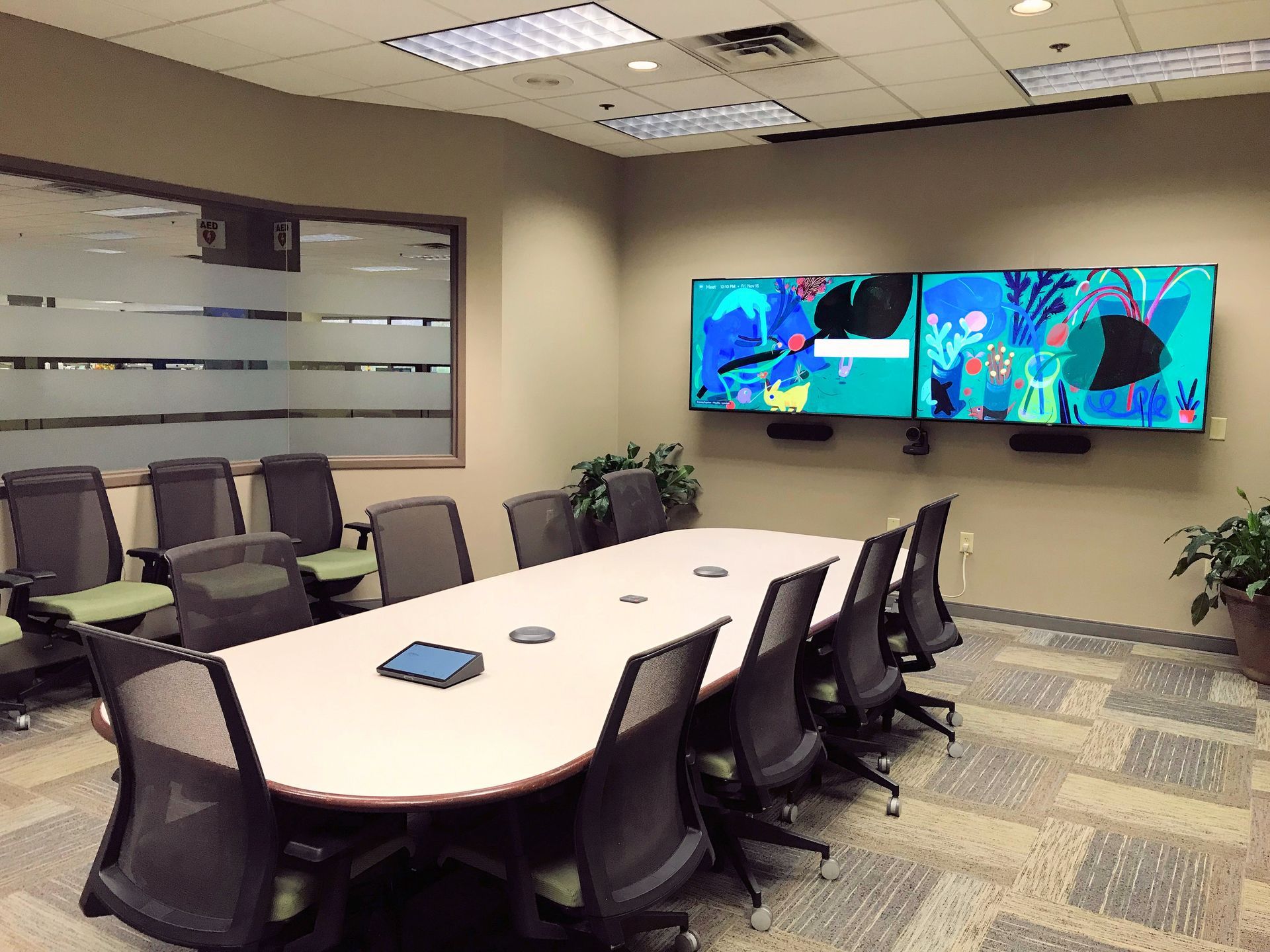 A conference room with a large table, multiple office chairs, and two wall-mounted screens displaying abstract art.