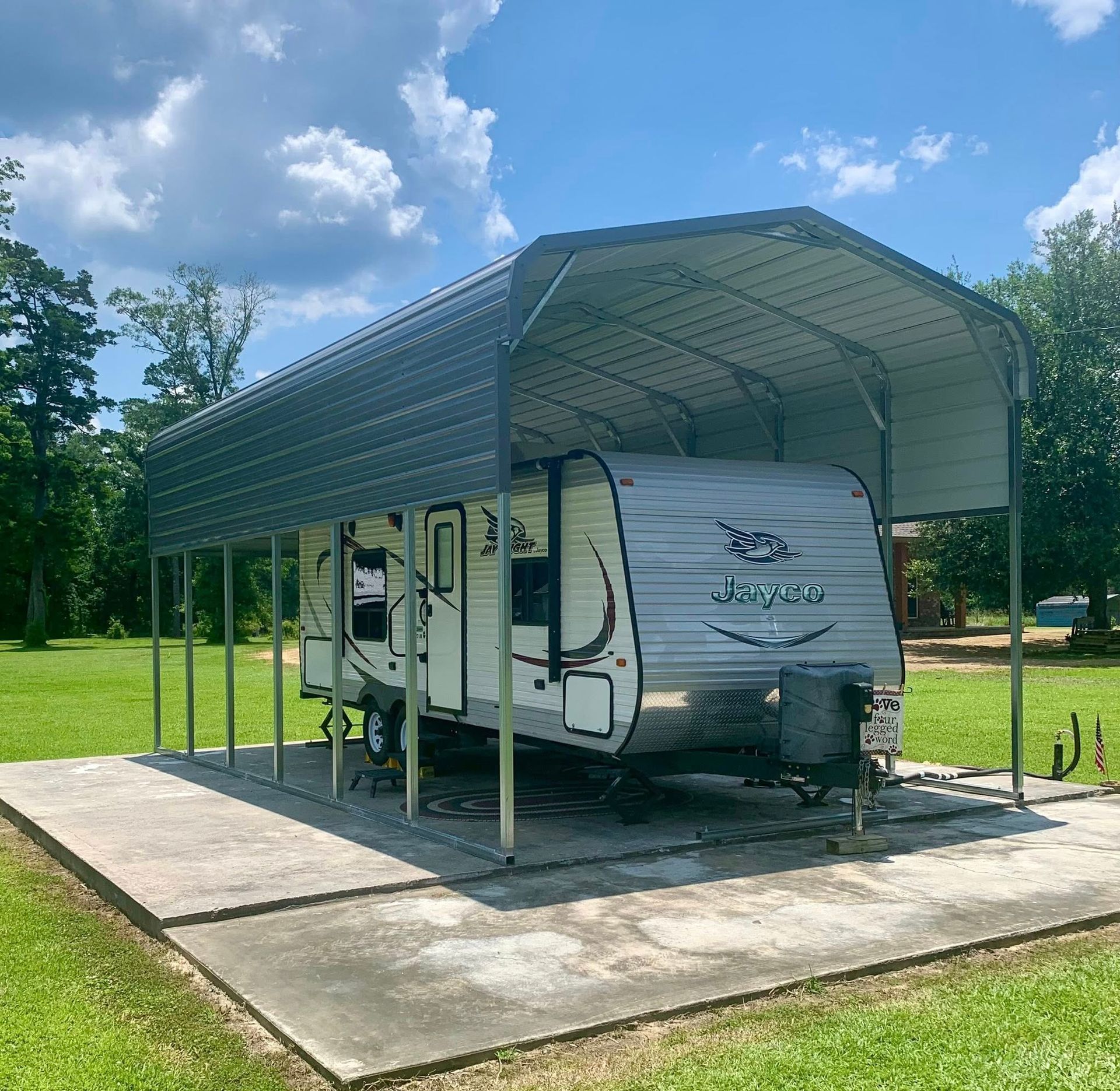 Gray camper parked under a metal carport on a concrete pad, outdoors on a sunny day.