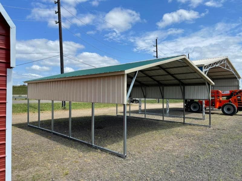 A carport with a green roof is sitting in a gravel lot next to a red barn.