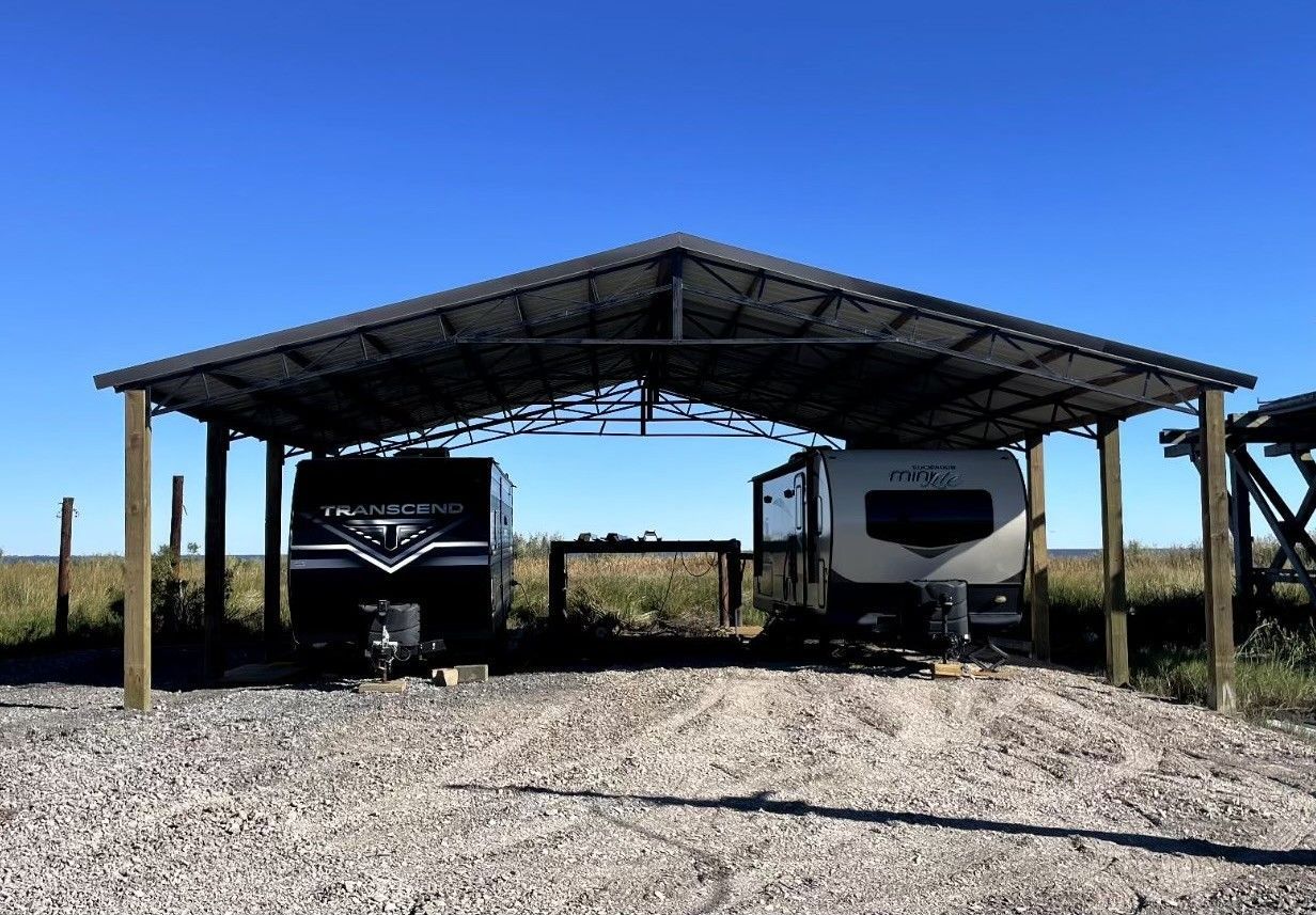 Two rvs are parked under a roof in a gravel lot.