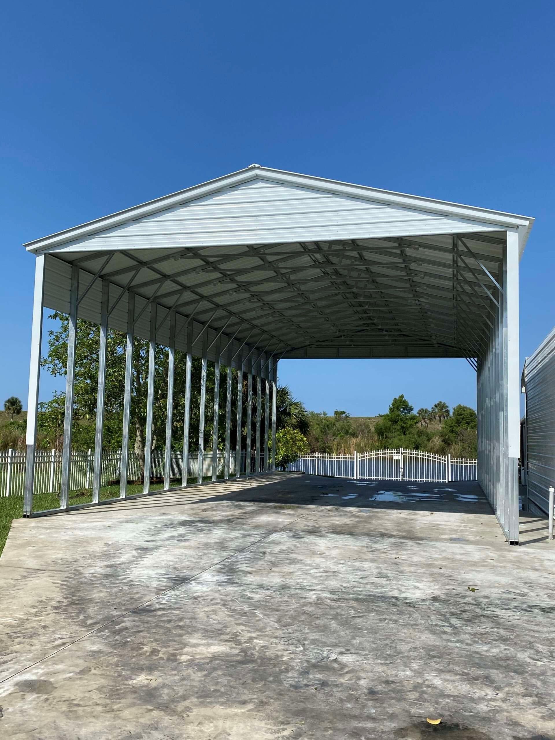 White metal carport with open sides, sits on a concrete pad, blue sky backdrop.