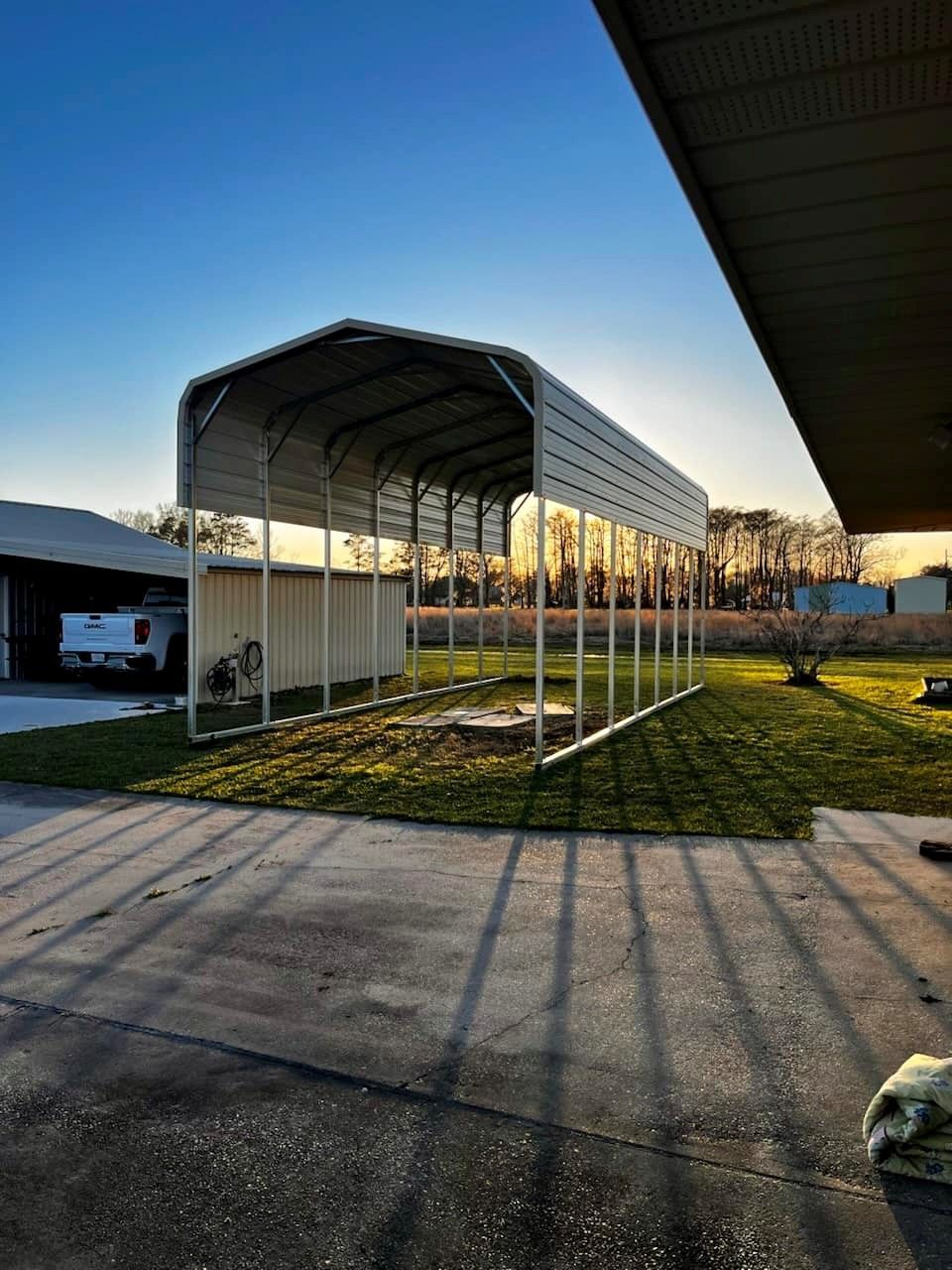 Metal carport in yard; white frame, tan roof, sunlit green grass and blue sky.