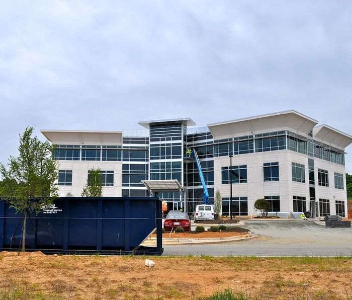 Three-story office building under construction; blue lift, dumpster, and car in front. Overcast sky.