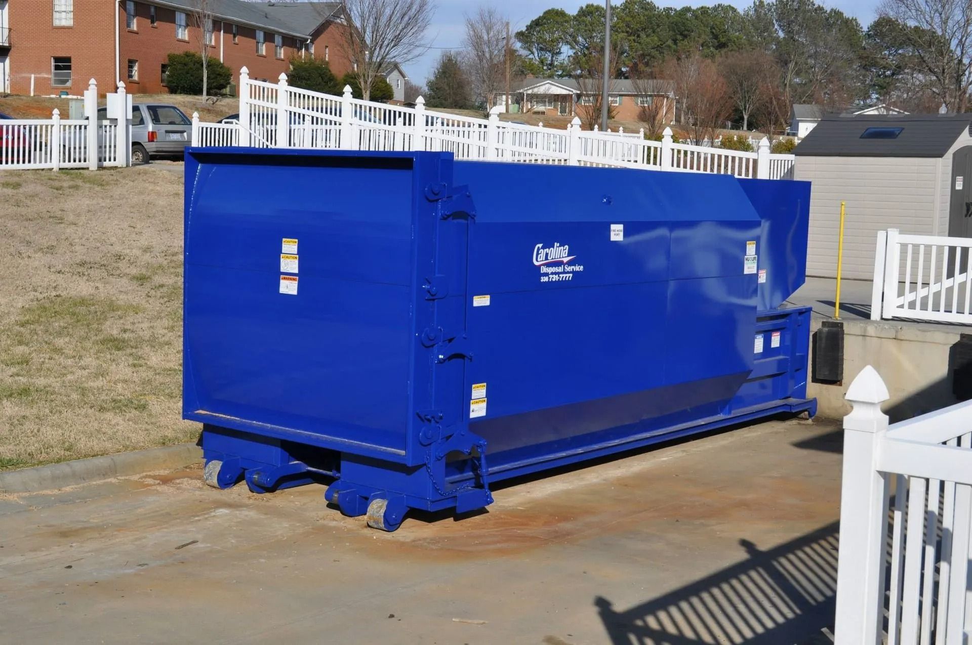 Large blue commercial dumpster, parked outdoors near a fence and buildings.