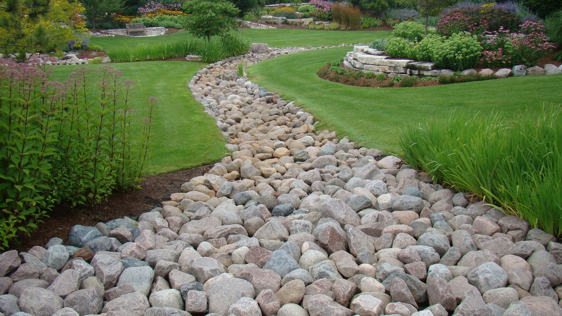 A dry creek bed made of gray river stones winds through a lush green lawn in a landscaped backyard.