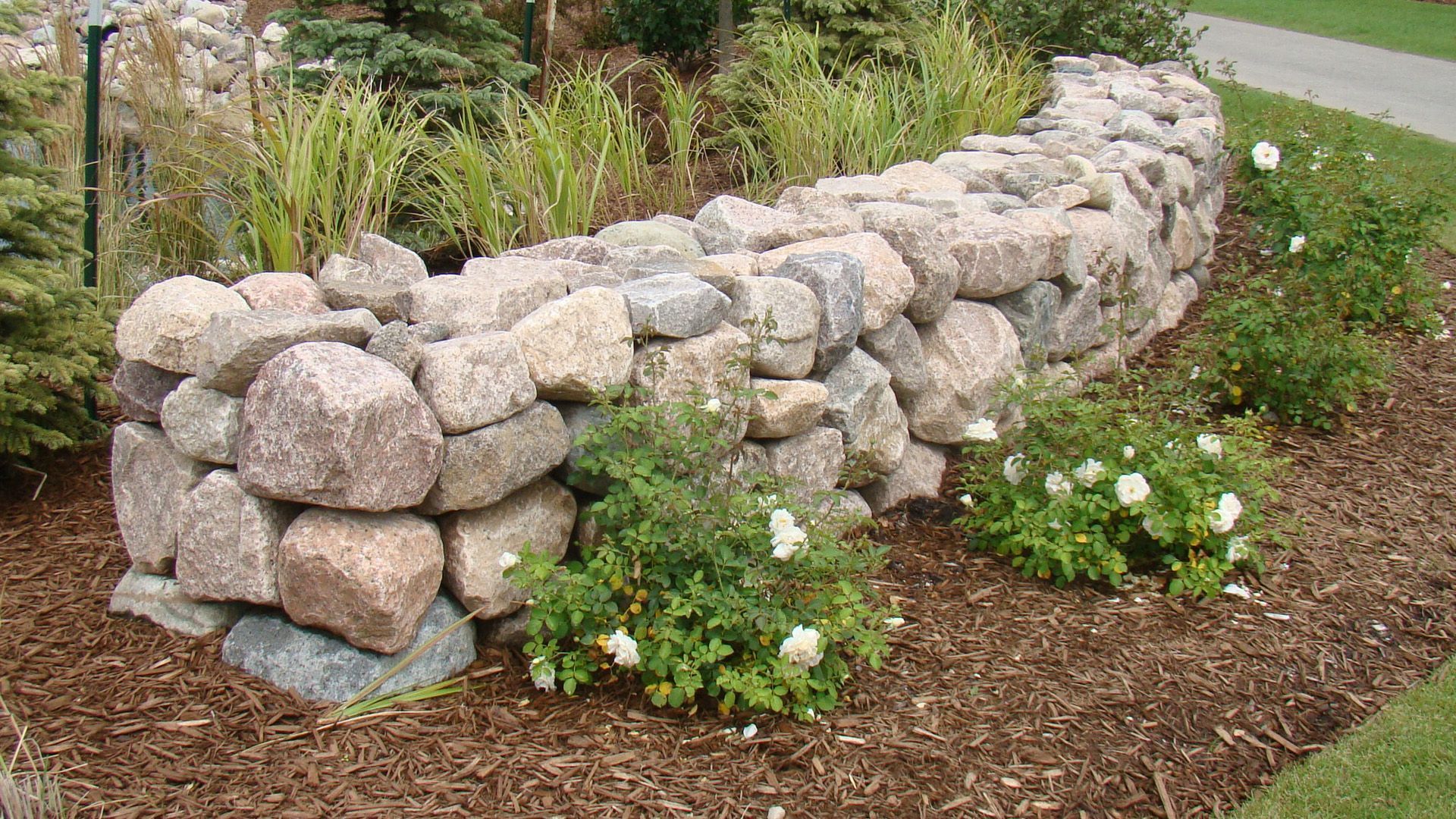 Curved stone garden wall along a mulched path, bordered by green plants and white flowers.