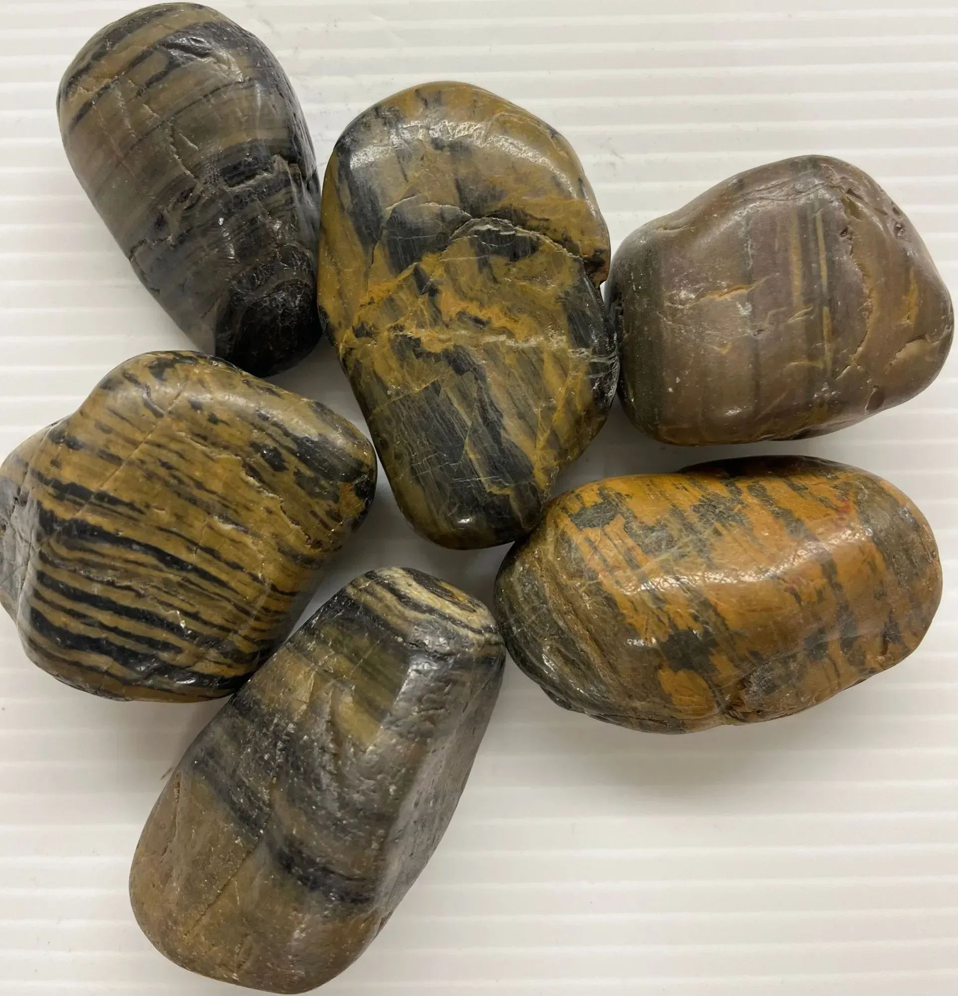 Six polished, tumbled stones with earthy brown and black striped patterns, arranged on a white ribbed surface.