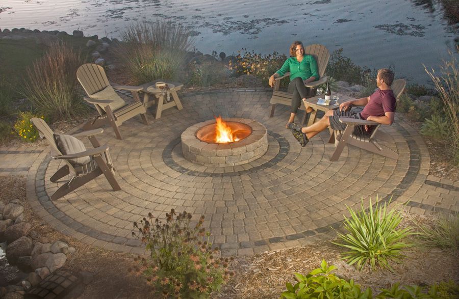 Two people sit in chairs around a circular stone fire pit on a patio overlooking a body of water at dusk.