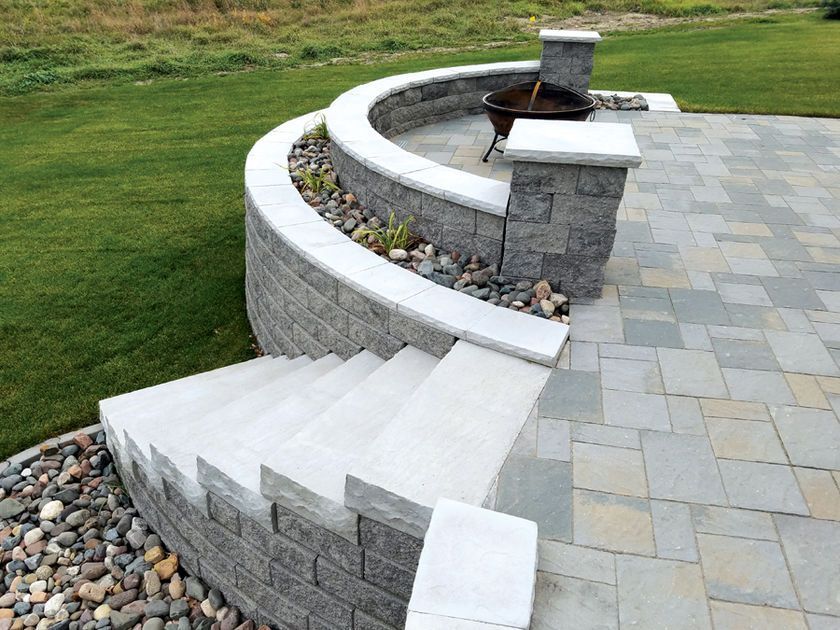 A curved stone retaining wall with integrated steps, a fire pit area, and a gray stone patio beside a green lawn.