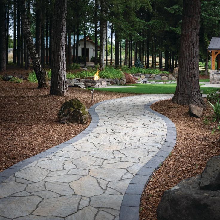 A stone pathway curves through a wooded backyard toward a stone fire pit area with lush green grass and tall trees.