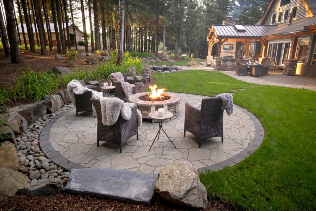 A circular stone patio with a fire pit and armchairs, set in a backyard near a house and forest.