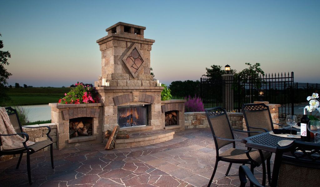 A stone outdoor fireplace with an active fire on a patio overlooking a field at sunset, with dining chairs nearby.