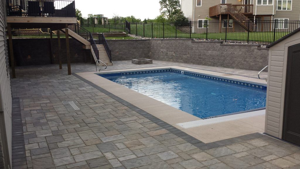 A backyard with a rectangular inground pool, gray stone pavers, a stone retaining wall, and a wooden deck.