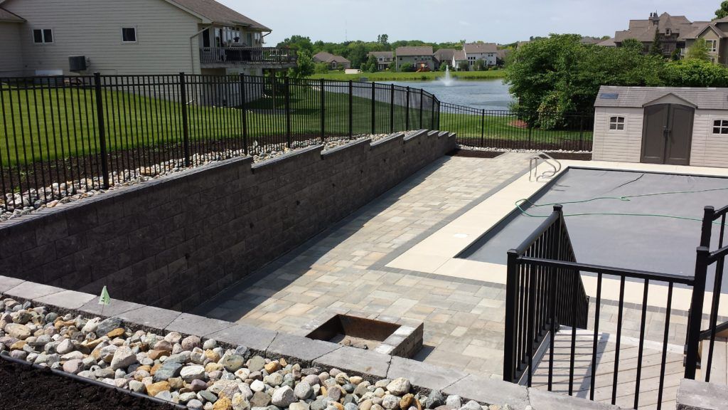 A backyard pool with a gray cover, stone patio, fire pit, and a dark stone retaining wall overlooking a lake.