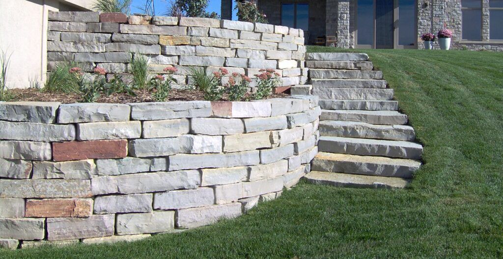 A tiered stone retaining wall beside a set of stone steps leading to a house, surrounded by a green lawn.