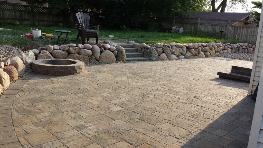 A paved stone patio with a circular fire pit and a tiered stone retaining wall leading to a grassy backyard.
