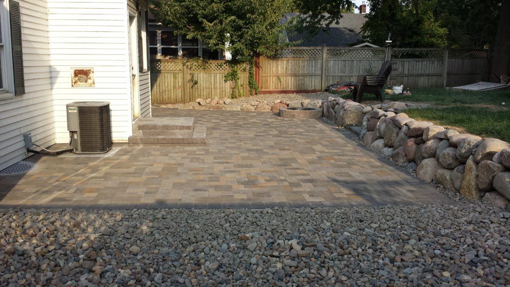 A stone paver patio sits beside a white house, bordered by a large fieldstone retaining wall and a gravel foreground.