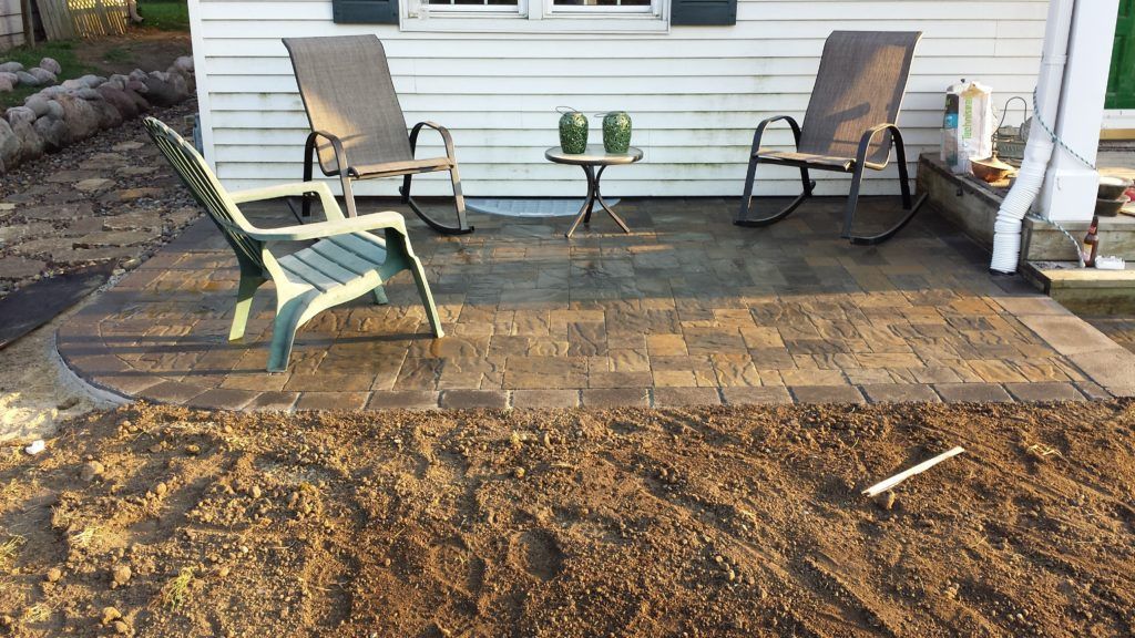 A patio made of stone pavers with two rocking chairs and one garden chair, set against a white house with bare soil in front.