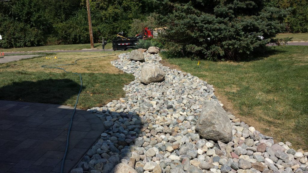 A winding dry creek bed made of smooth gray stones and large boulders, bordered by grass and a paved patio edge.