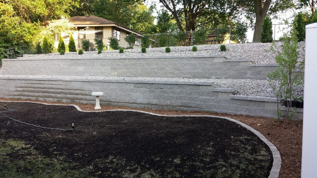 A terraced backyard with grey stone retaining walls, gravel landscaping levels, dark mulch garden beds, and a small tree.