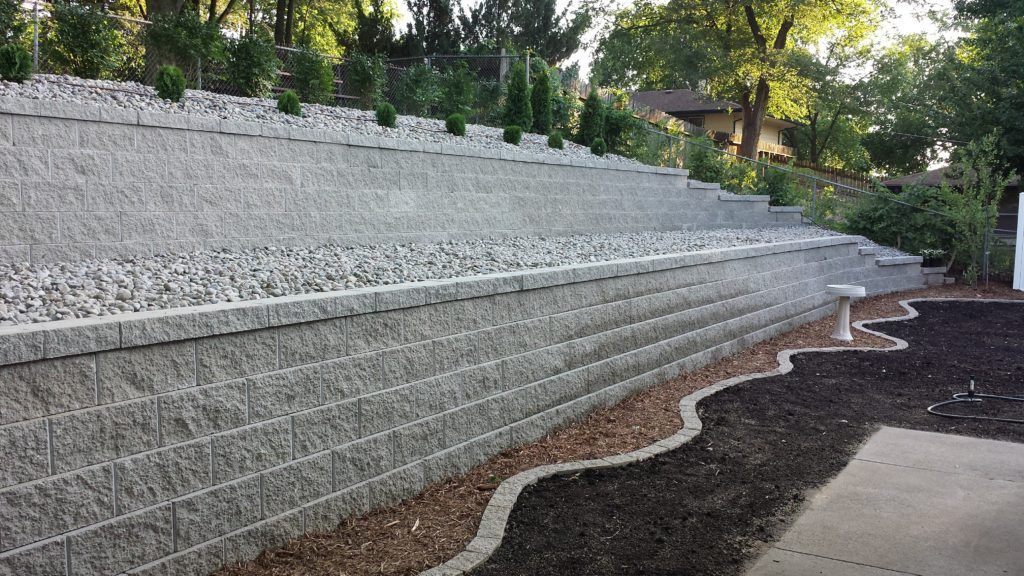 A multi-tiered gray retaining wall with gravel landscaping and small trees, bordering a mulched garden bed and patio.