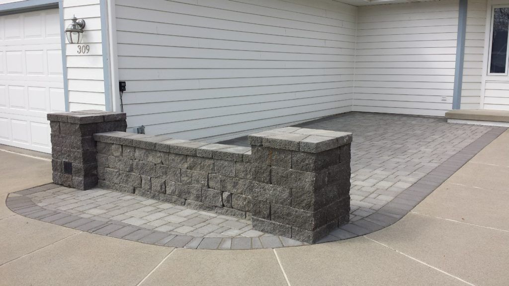 A stone low wall with two pillars bordering a grey paver patio next to a white house.