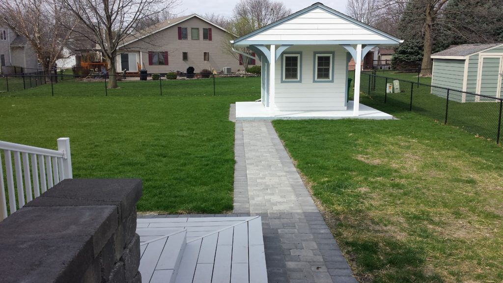A stone path leads across a grassy lawn to a small, white structure with a peaked roof, viewed from a wooden deck.