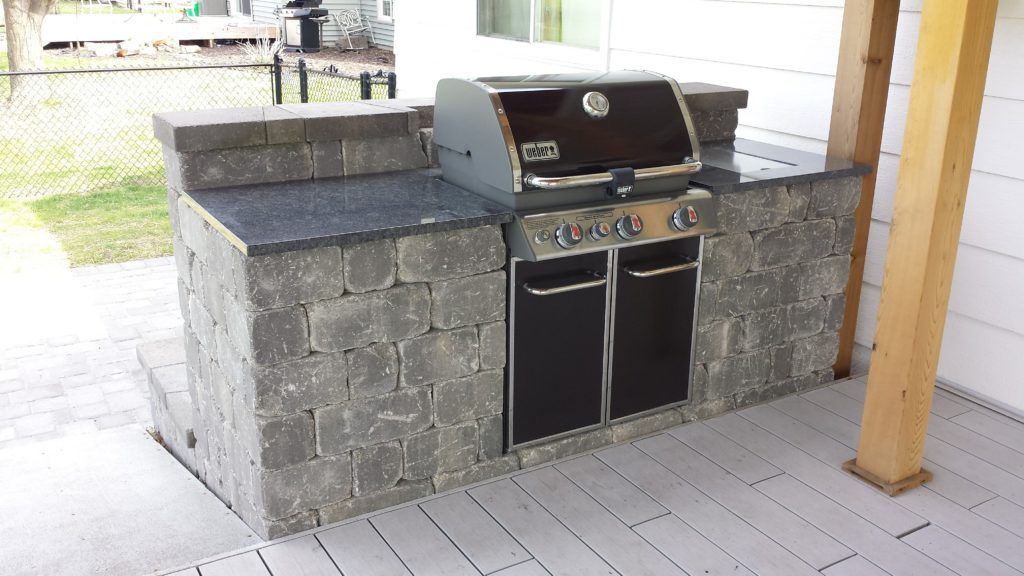 An outdoor kitchen featuring a gray stone grill station with a dark granite countertop and a stainless steel gas grill.