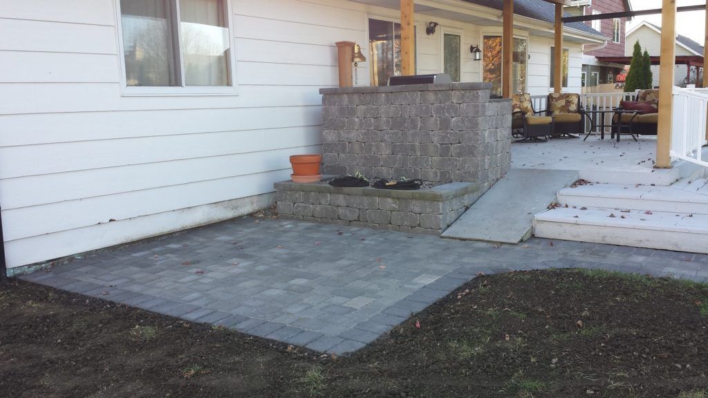 A stone patio with a built-in outdoor fireplace and a ramp leading up to a wooden-covered porch area.