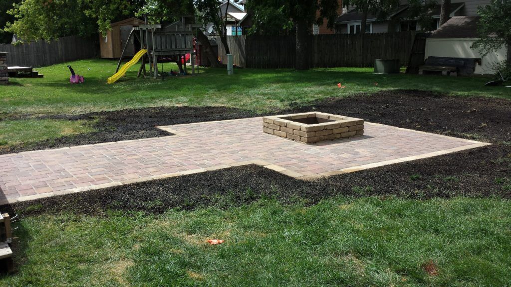 A stone fire pit sits in the center of a newly installed rectangular paver patio, surrounded by fresh dark mulch and grass.