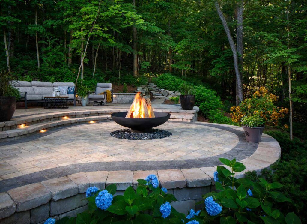 A stone patio featuring a glowing fire bowl, surrounding seating, and blue hydrangea flowers in a lush forest setting.