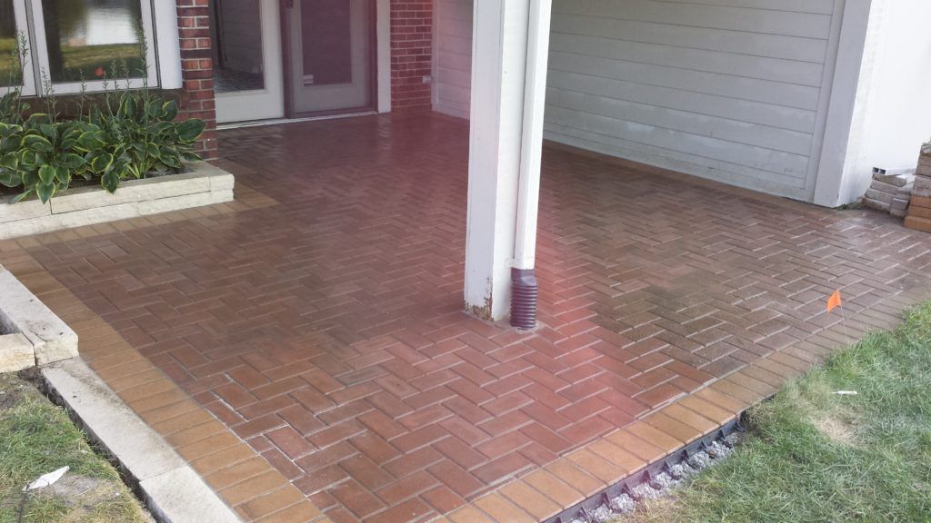 A red brick patio with a herringbone pattern, framed by a tan border, in front of a house entrance and garage.